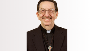 Smiling deacon in glasses and black suit with cross pendant, posed against a plain white background.