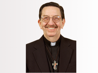 Smiling deacon in glasses and black suit with cross pendant, posed against a plain white background.