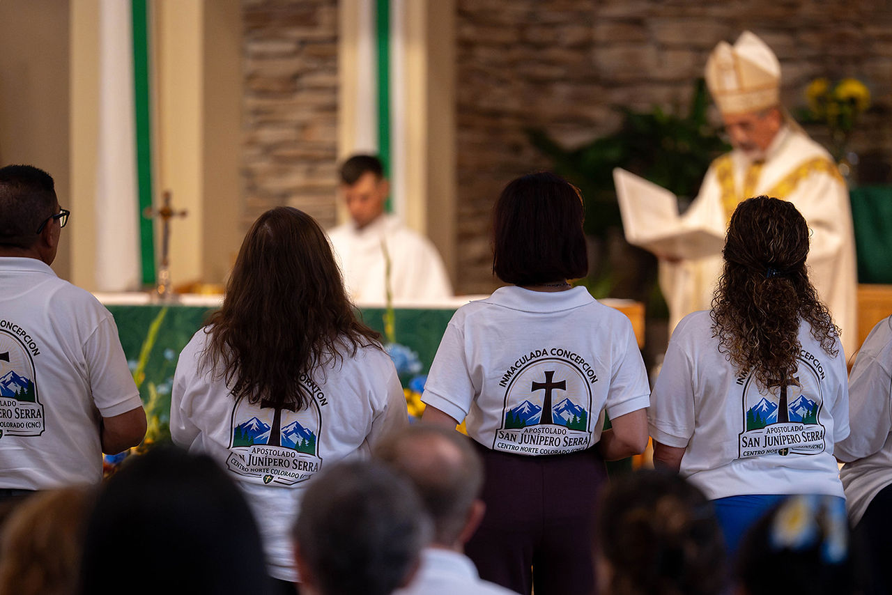 People in white San Junípero Serra shirts stand at a church Mass while a priest reads at the altar with a cross and green cloth.