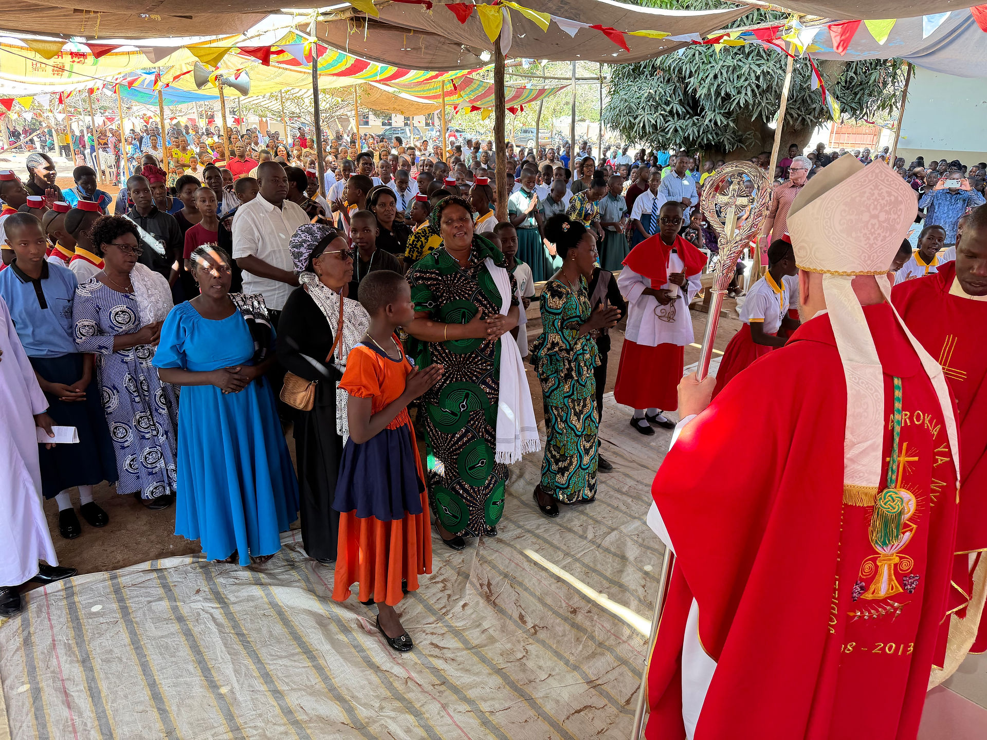 Crowd gathered under colorful canopies at an outdoor church service, facing red-robed clergy with a ceremonial staff and cross.