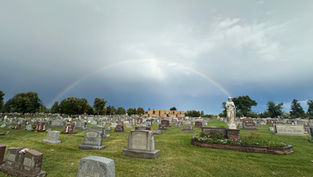 Rainbow over a cemetery with many gravestones, a white statue, and a cloudy sky after rain.