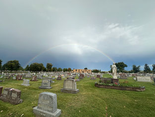 Rainbow over a cemetery with many gravestones, a white statue, and a cloudy sky after rain.