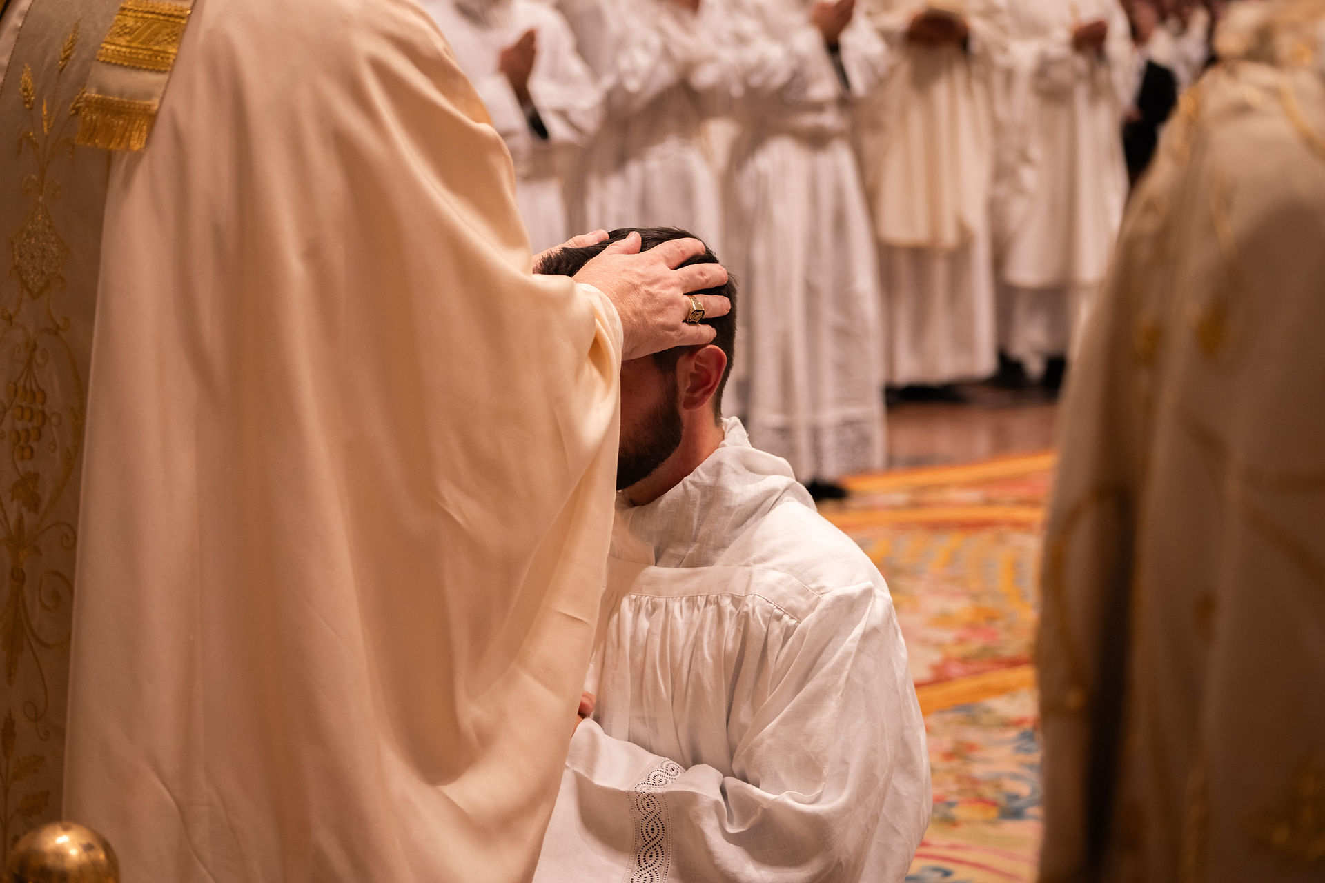 Bishop lays hands on kneeling man during solemn church ordination ceremony, with white-robed clergy in background
