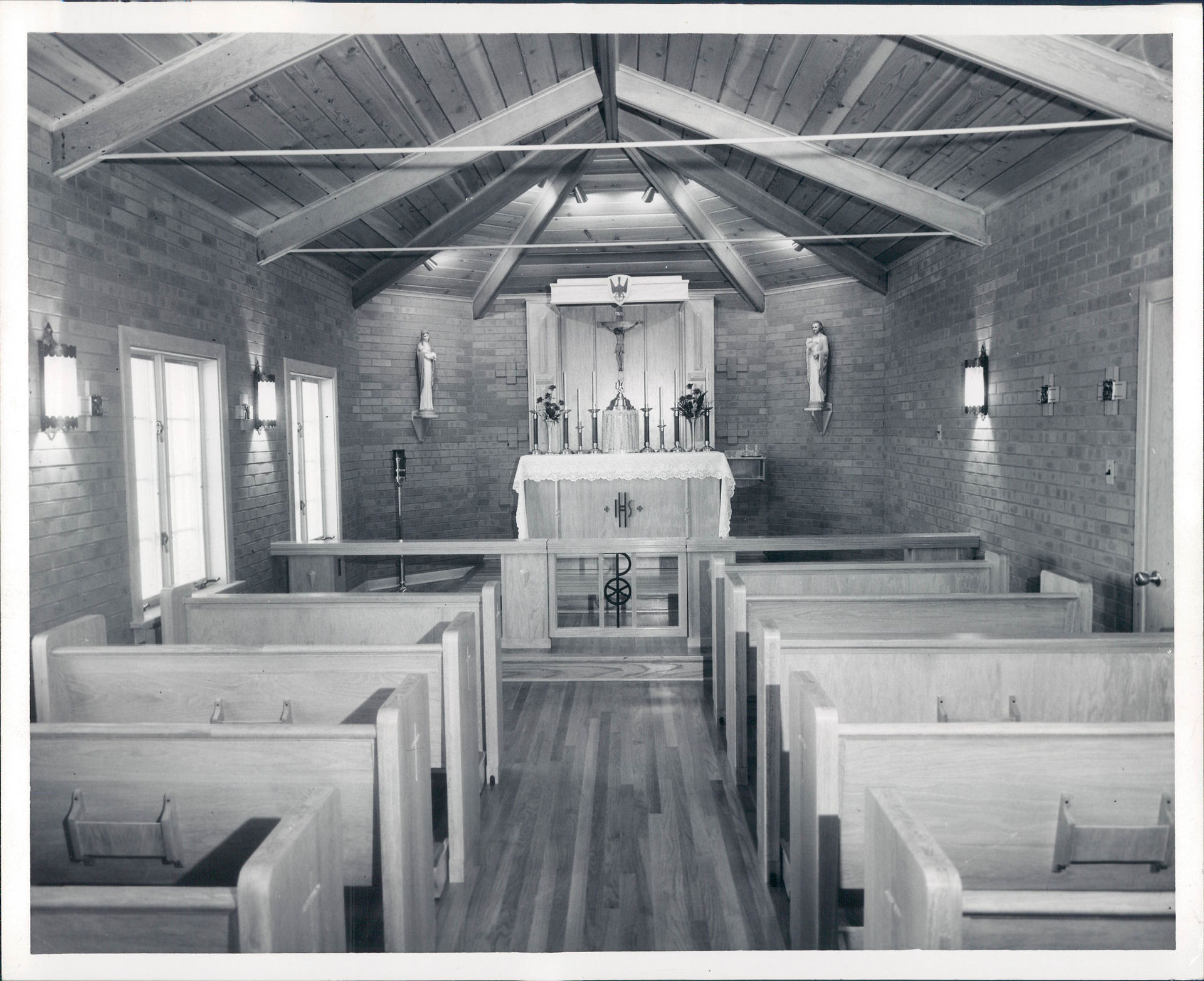 Empty chapel with wooden pews, crucifix and altar at the front, brick walls, slanted beam ceiling, and soft window light.