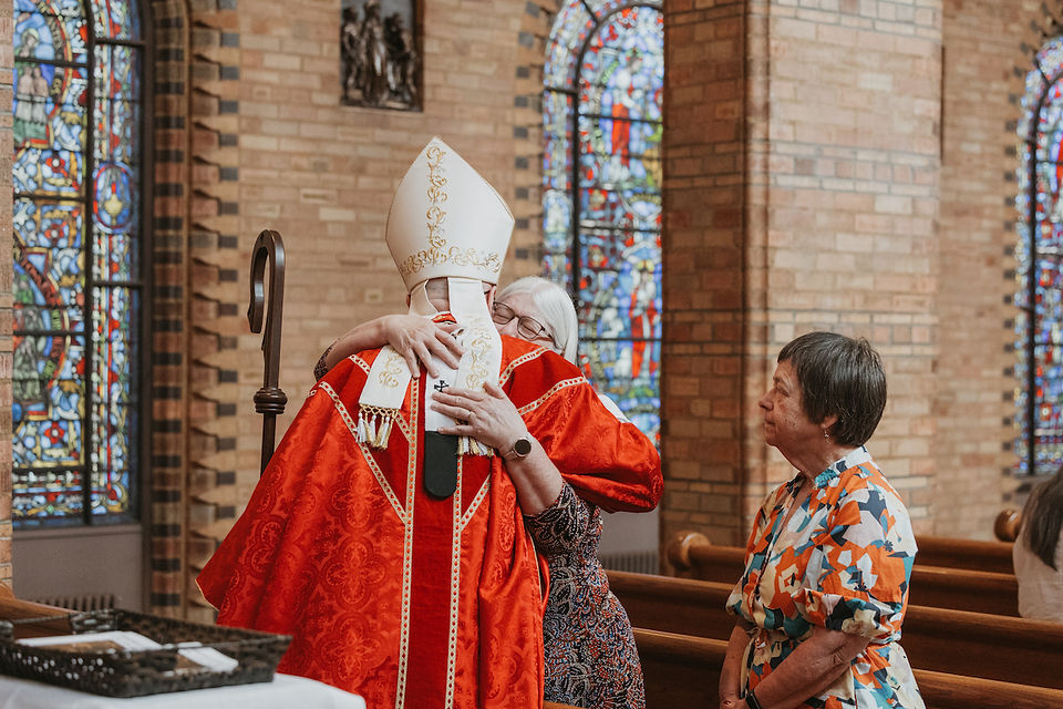 Bishop in red vestments hugs an elderly woman in a church with stained-glass windows; another woman watches warmly.
