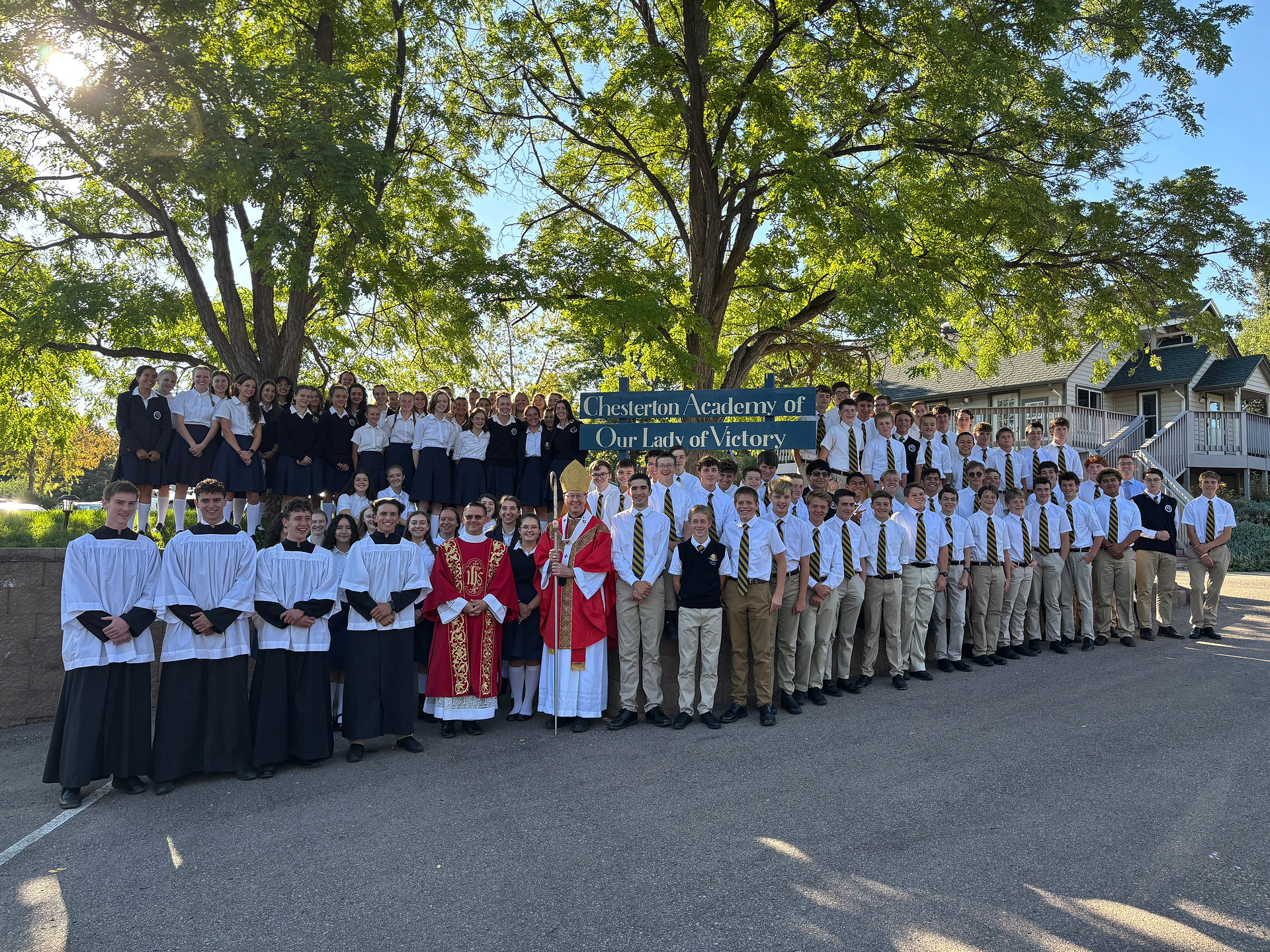 Large group of uniformed students and clergy pose outdoors by a sign reading Chesterton Academy of Our Lady of Victory.