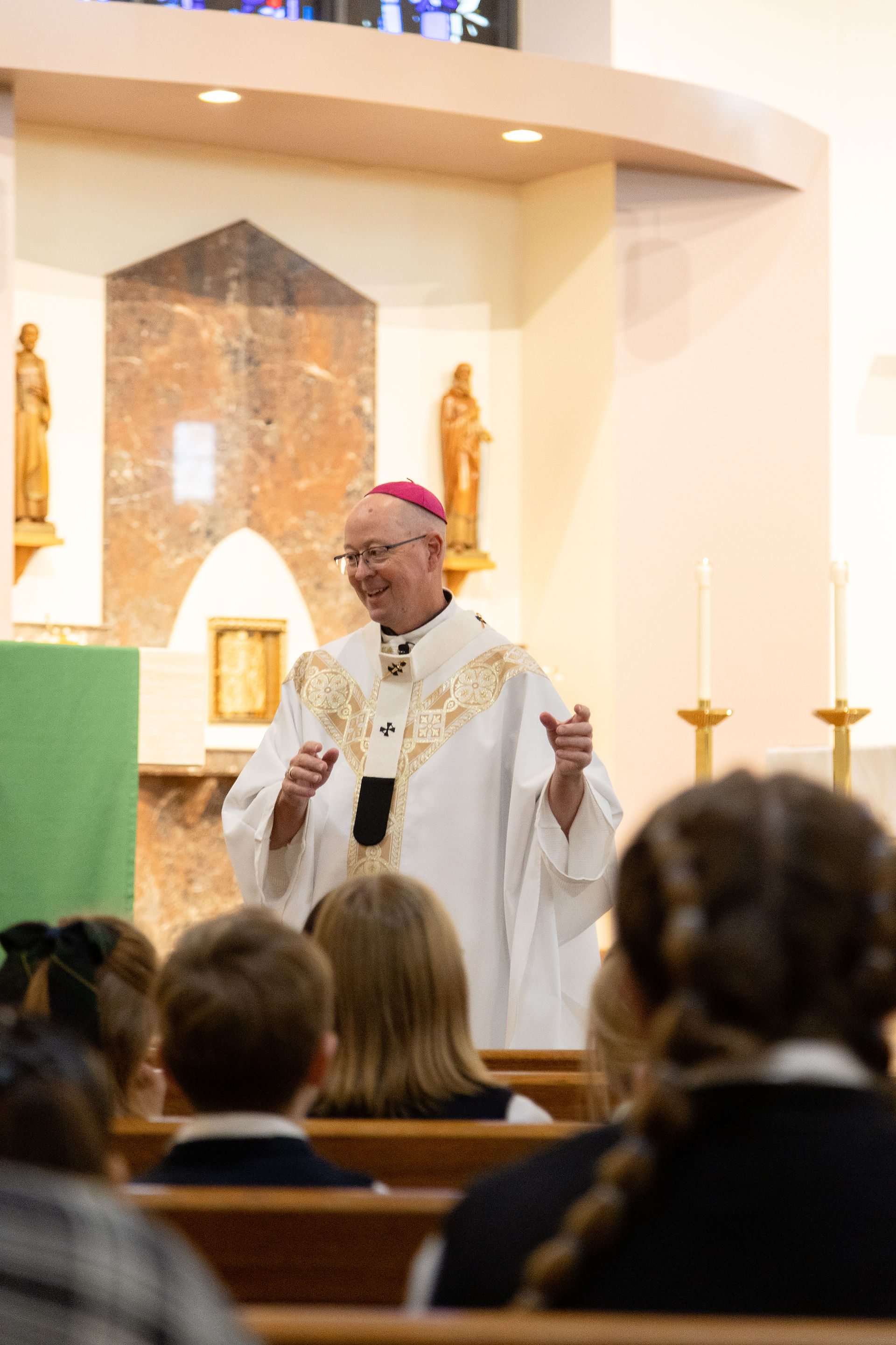 Smiling bishop in white vestments and magenta skullcap speaks to seated congregation in a church, with altar and statues behind him.