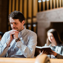 Man praying in a church pew, with two women behind him—one reading a book—warm light and a solemn mood