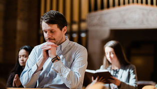 Man praying in a church pew, with two women behind him—one reading a book—warm light and a solemn mood