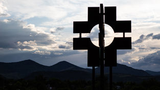 Silhouetted geometric sign against cloudy dusk sky, with mountains and trees in the background.