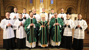 Smiling Catholic clergy and seminarians pose in green and white vestments inside a church with stained glass and altar.