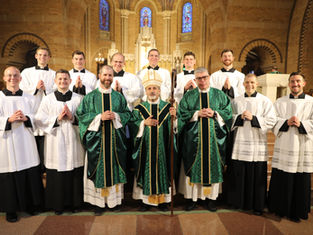 Smiling Catholic clergy and seminarians pose in green and white vestments inside a church with stained glass and altar.