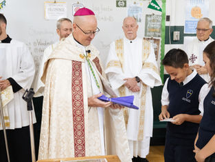 Bishop in ornate vestments receives a gift wrapped in purple cloth from two schoolchildren, with priests behind him in a classroom with posters.