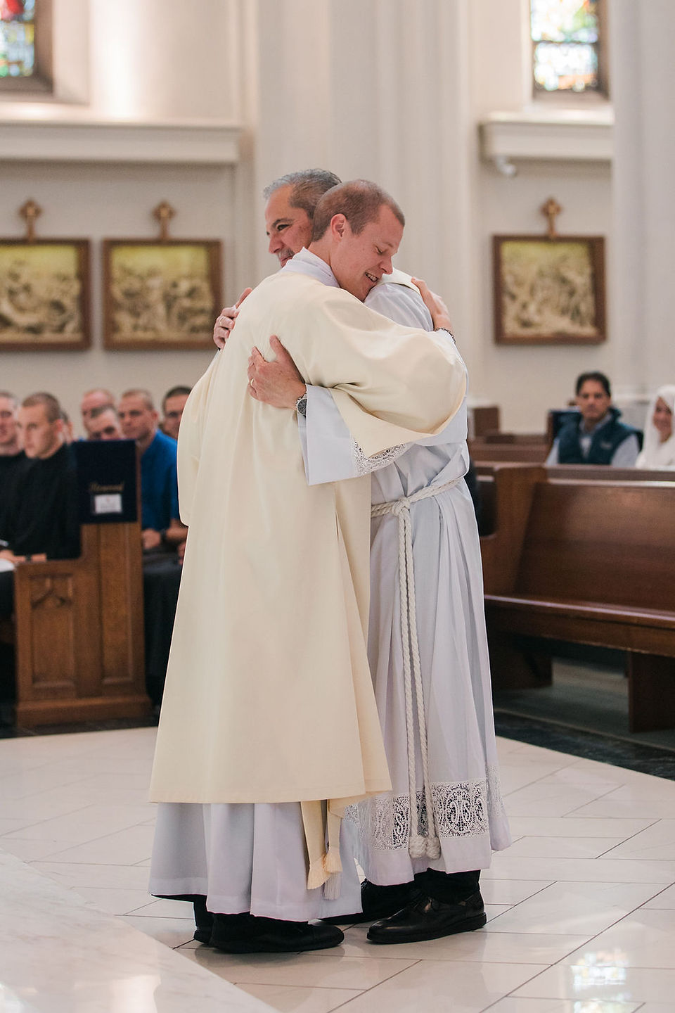 Two deacons in white robes hug warmly inside a church, with seated parishioners and stained-glass windows in the background.