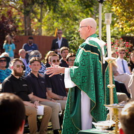 Bald priest in green vestments speaks to a seated outdoor congregation in a sunny garden ceremony.