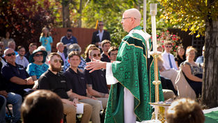 Bald priest in green vestments speaks to a seated outdoor congregation in a sunny garden ceremony.