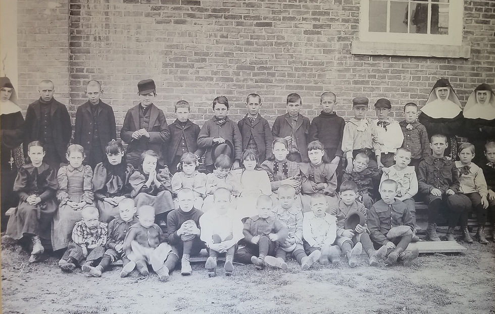 Black-and-white class photo of children and a few nuns posing solemnly against a brick wall outside a school.