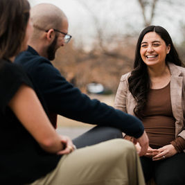 Three people sit outdoors talking; a smiling woman in a plaid blazer faces a couple in the foreground on a cloudy day.