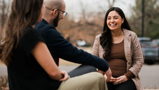 Three people sit outdoors talking; a smiling woman in a plaid blazer faces a couple in the foreground on a cloudy day.
