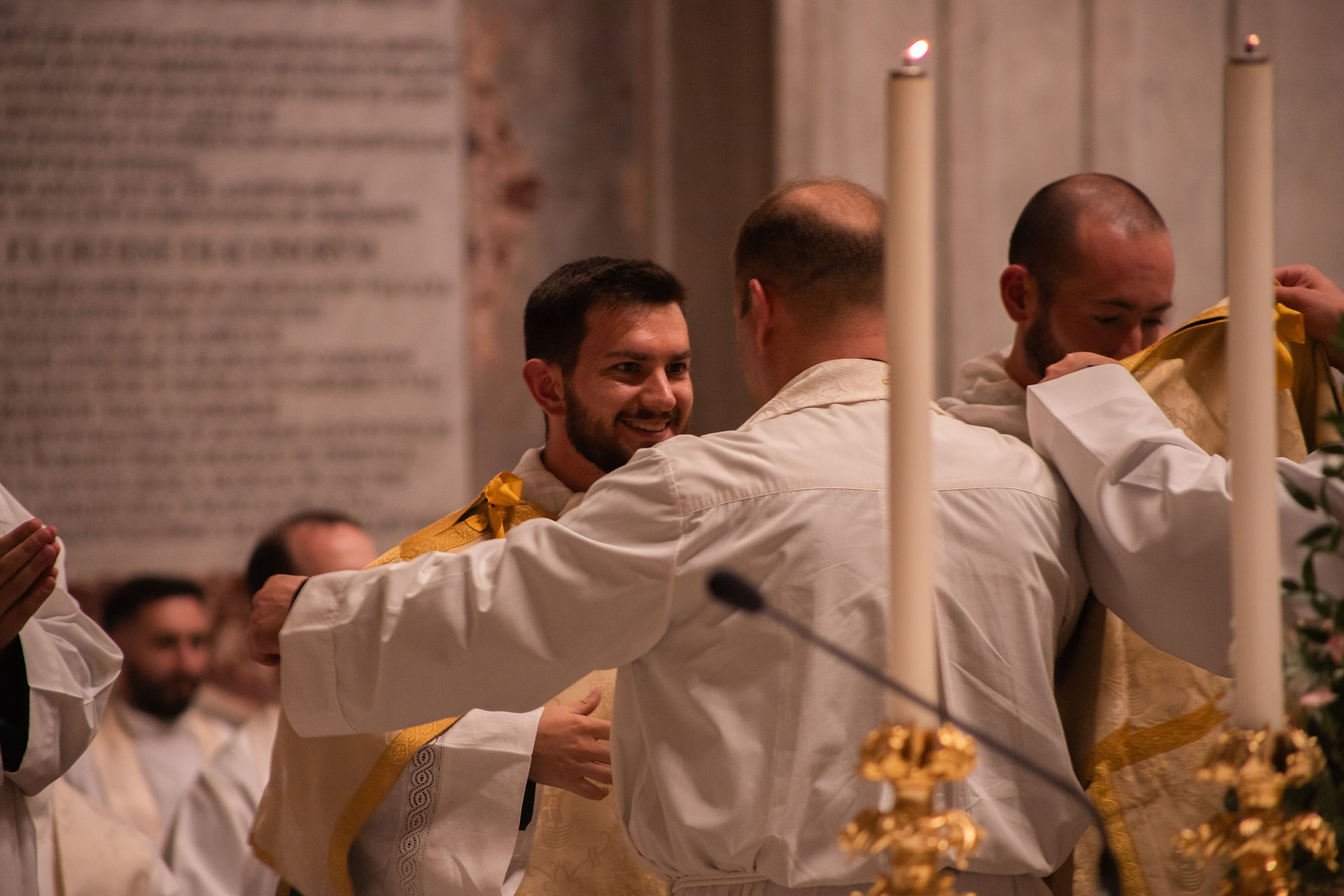 Priests in white and gold vestments embrace during a church ceremony, with candles glowing in the foreground.