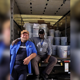 Two smiling men sit at the back of a truck loaded with gray paint buckets and boxes, in a warehouse-like setting.