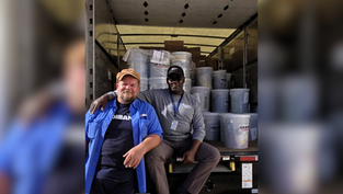 Two smiling men sit at the back of a truck loaded with gray paint buckets and boxes, in a warehouse-like setting.