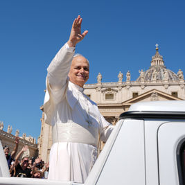 Pope in white waves from an open vehicle outside St. Peter’s Basilica as crowds cheer under a clear blue sky.