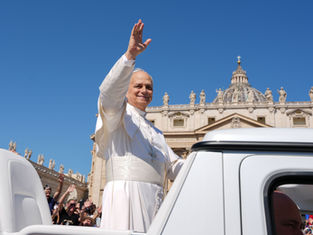 Pope in white waves from an open vehicle outside St. Peter’s Basilica as crowds cheer under a clear blue sky.