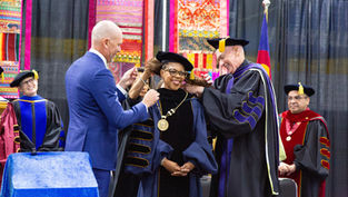 Officials in academic regalia place a gold medallion on a smiling woman at a graduation ceremony with colorful textile backdrops.