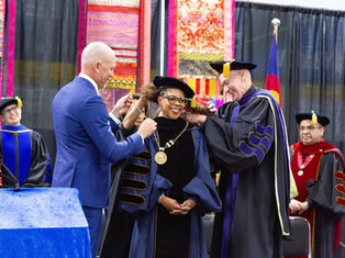 Officials in academic regalia place a gold medallion on a smiling woman at a graduation ceremony with colorful textile backdrops.
