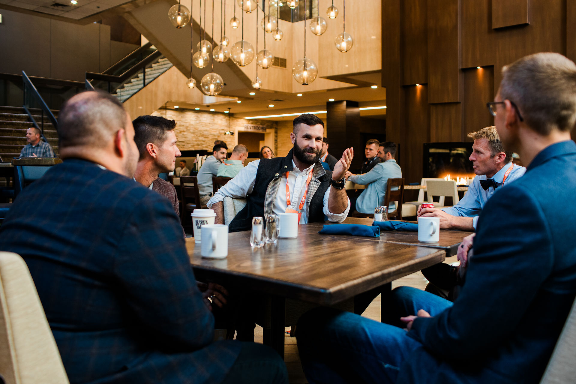 Men in suits chat around a wooden table in a warm hotel lounge with hanging globe lights and a visible RESTROOMS sign.