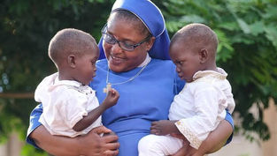 Smiling nun in blue holds two young children outdoors, looking at a small pendant, with green foliage behind them.
