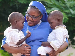 Smiling nun in blue holds two young children outdoors, looking at a small pendant, with green foliage behind them.