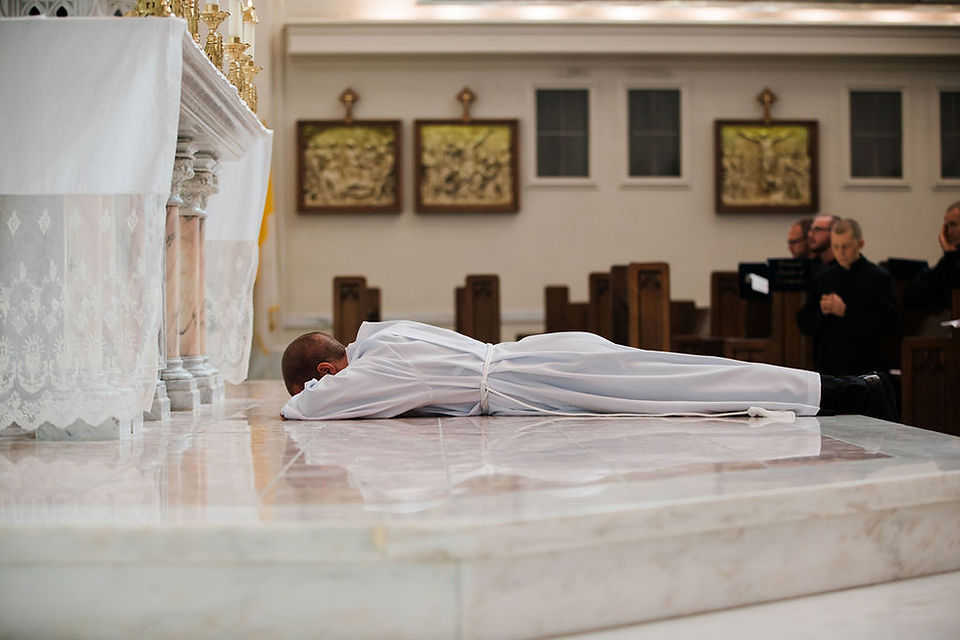 Man in white robe lies face down before a church altar, with clergy praying in the background.
