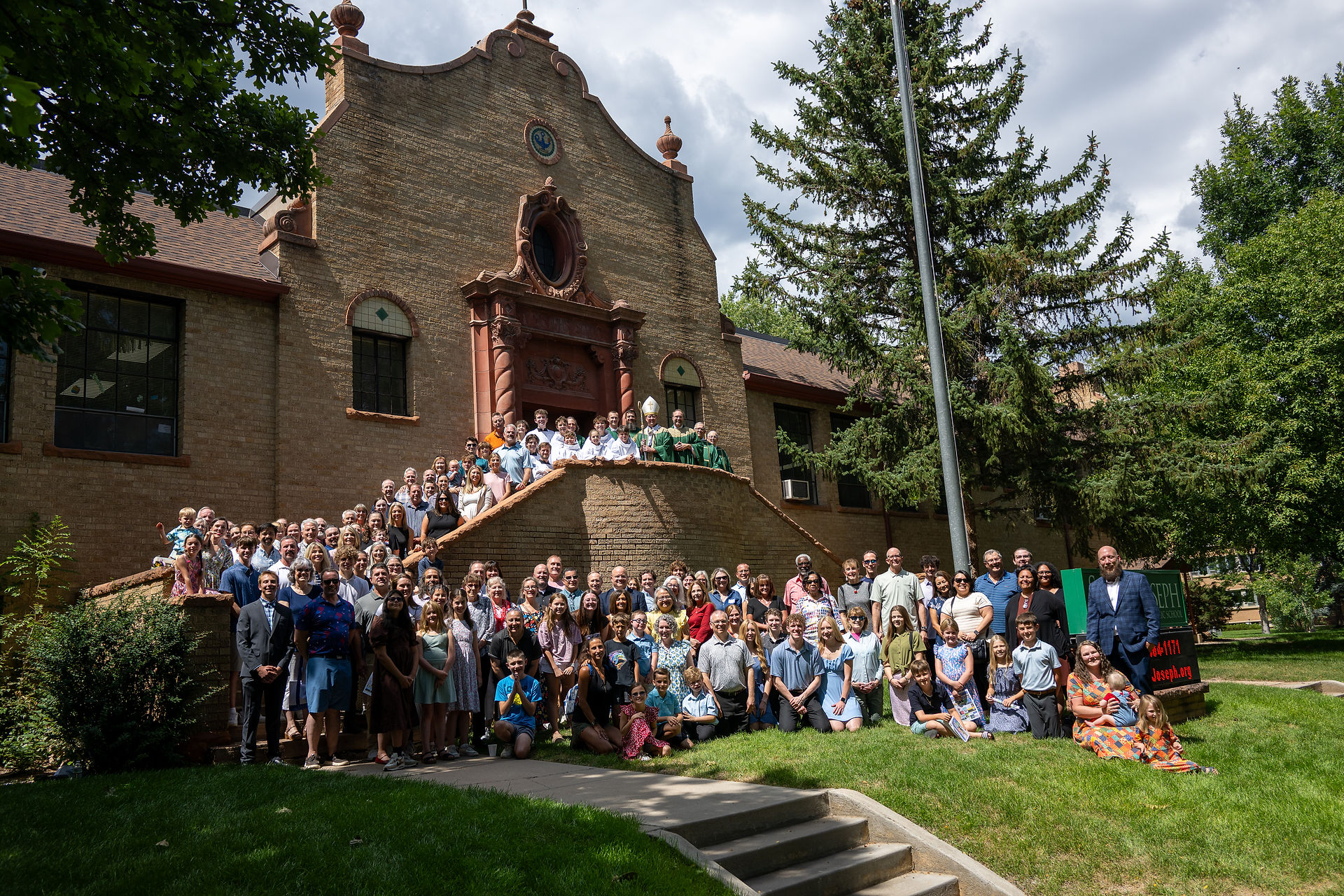Large group poses on steps outside a brick church, with clergy in green vestments and a green school sign at right.