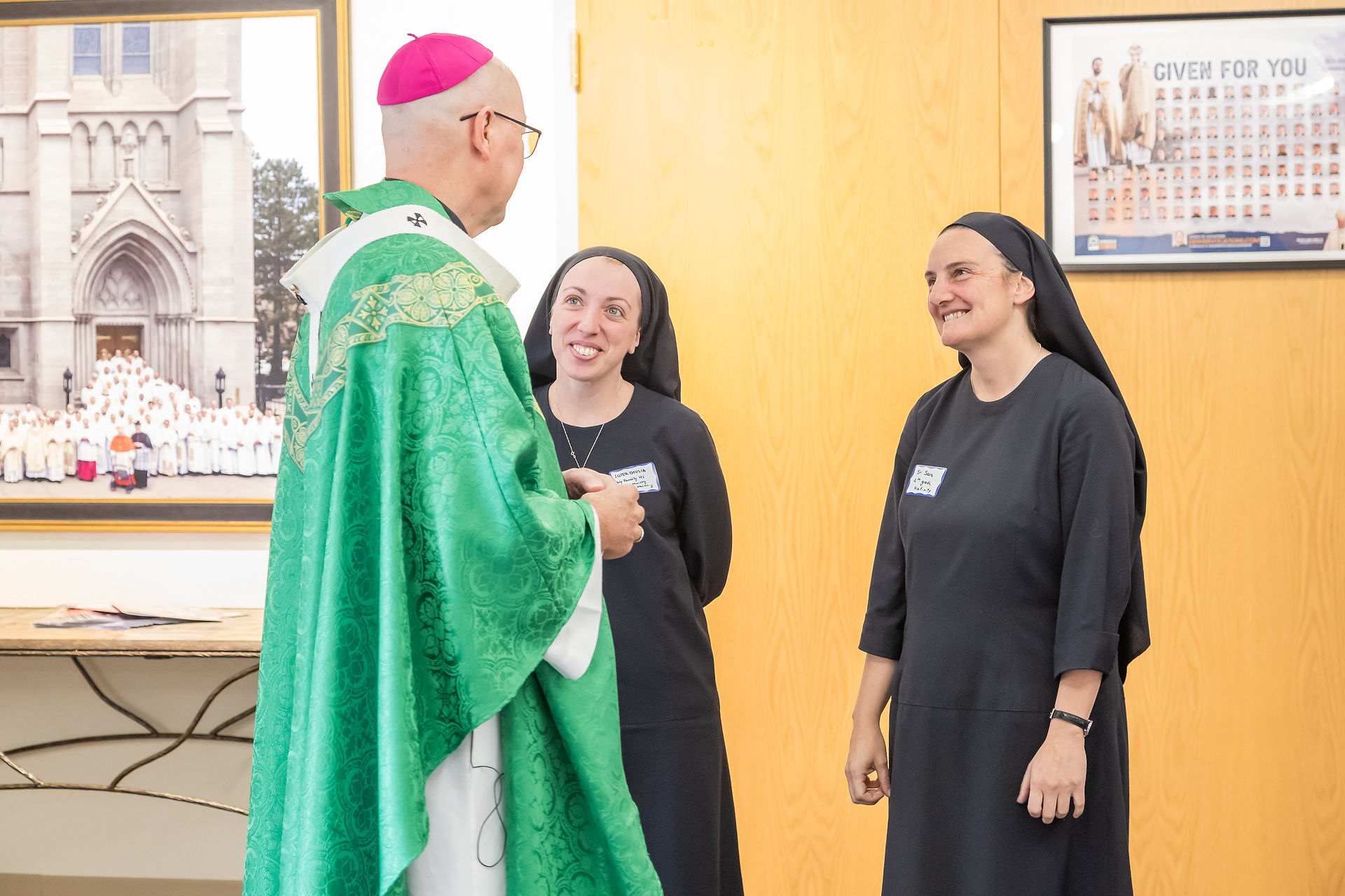 Bishop in green vestments chats with two smiling nuns in a bright church hallway; framed poster says GIVEN FOR YOU.