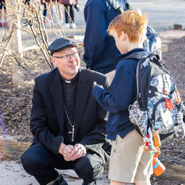 Priest kneels and talks to a boy with a backpack in a sunny outdoor park, with bare shrubs and other people behind.