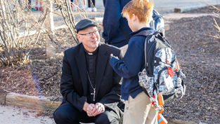 Priest kneels and talks to a boy with a backpack in a sunny outdoor park, with bare shrubs and other people behind.