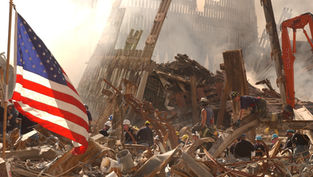 Rescue workers search rubble amid smoke and twisted steel, with a large U.S. flag in the foreground at a disaster site.