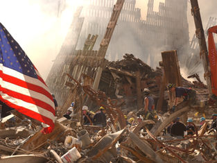 Rescue workers search rubble amid smoke and twisted steel, with a large U.S. flag in the foreground at a disaster site.
