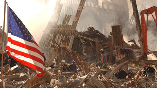 Rescue workers search rubble amid smoke and twisted steel, with a large U.S. flag in the foreground at a disaster site.