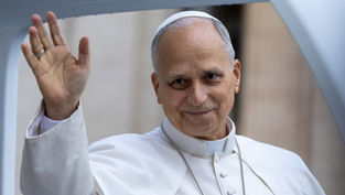 Pope Leo in white robes waves and smiles softly, with a blurred church-like background.