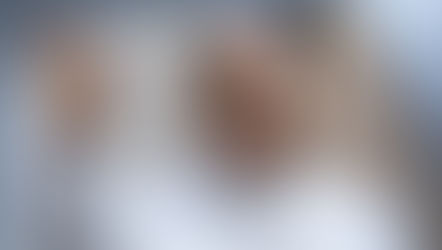 Pope Leo in white robes waves and smiles softly, with a blurred church-like background.