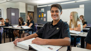 Smiling boy in black polo sits at a classroom desk with an open book, while classmates study behind him near a whiteboard.