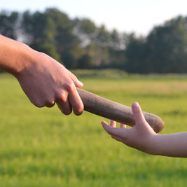 Adult hand passes a wooden baton to a child’s outstretched hand in a sunny grassy field with trees.