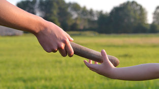 Adult hand passes a wooden baton to a child’s outstretched hand in a sunny grassy field with trees.
