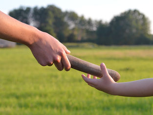 Adult hand passes a wooden baton to a child’s outstretched hand in a sunny grassy field with trees.