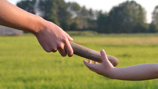 Adult hand passes a wooden baton to a child’s outstretched hand in a sunny grassy field with trees.