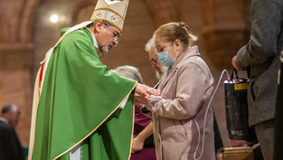 Bishop in green vestments blesses a masked woman during a church service.