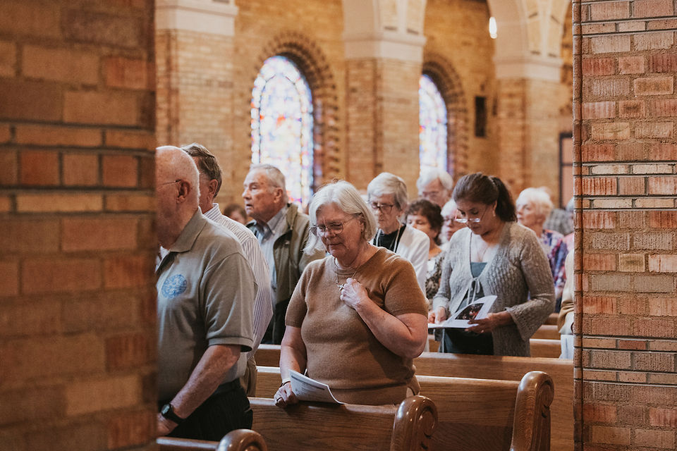 Elderly congregants stand in wooden church pews during a service, holding papers under stained glass windows in a quiet, solemn mood